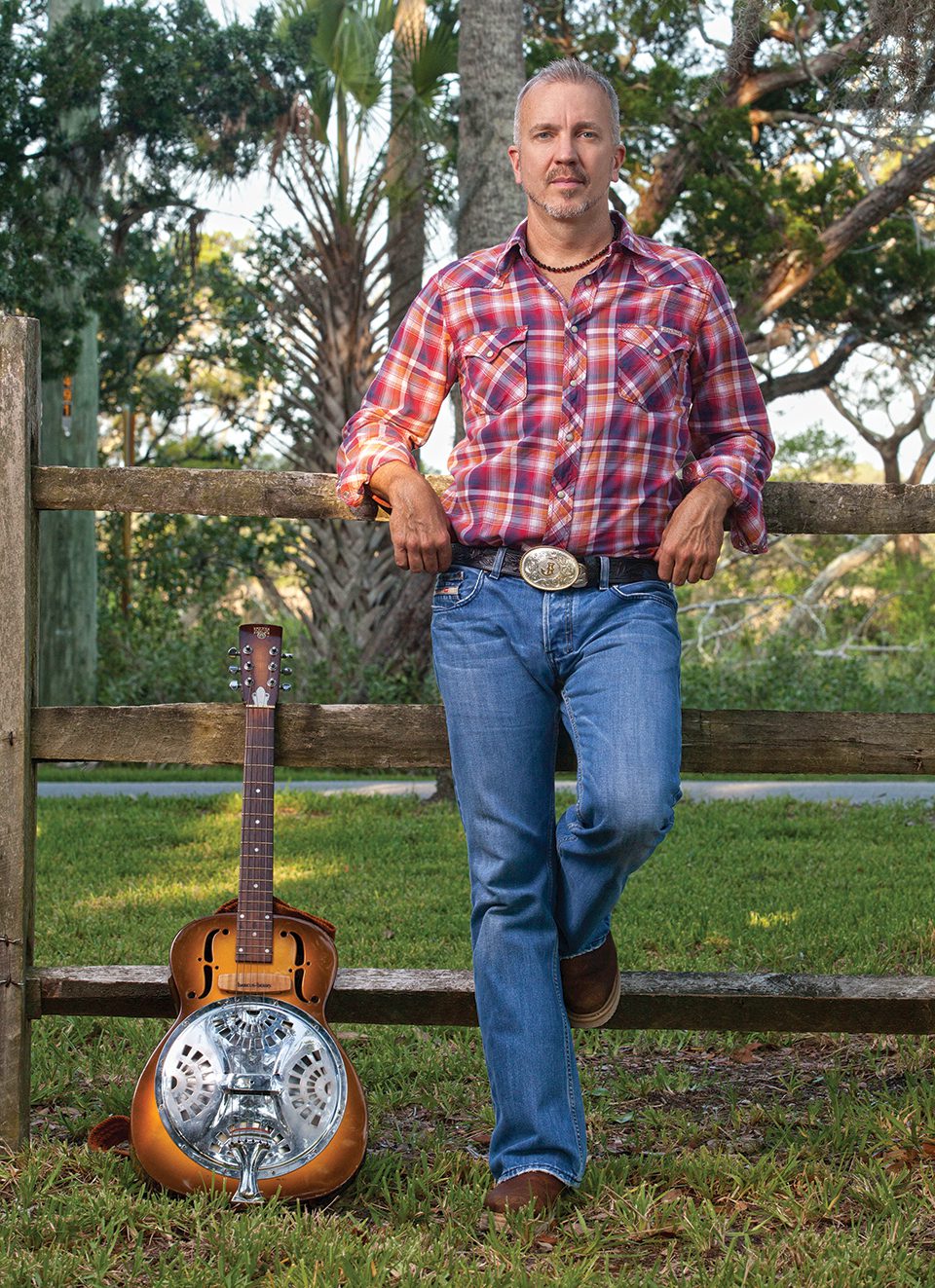 JJ Grey wears a red plaid shirt, a belt with a large buckle and jeans as he and his Dobro guitar rest against a wooden fence.