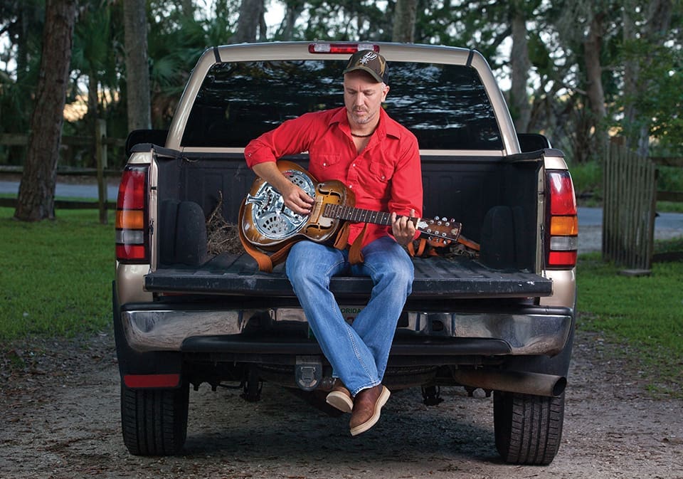 JJ Grey strums a Dobro guitar. He is in the truck bed and wears a red shirt and jeans.