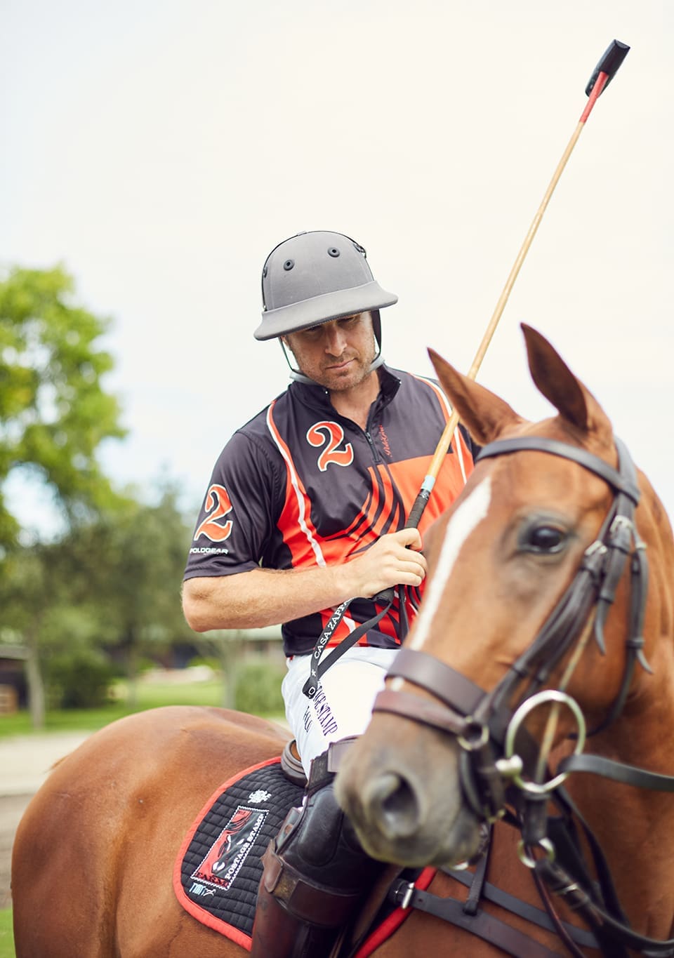 Brandon Phillips sits on his horse Honey Badger who is out of focus glancing slyly at the camera. Phillips wears a jersey and helmet and holds his mallet.