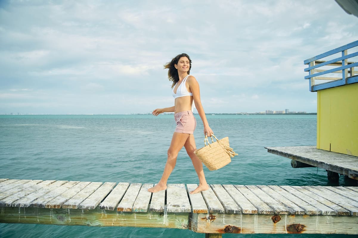 Stiltsville: Stilted, Jilted & Re-builted 5 Ashley struts down a Stiltsville home's dock. She is barefoot and wears a white bikini top and blush pink shorts. She carriers a jute tote bag. Water stretches out behind her and the skyline is visible in the distance.