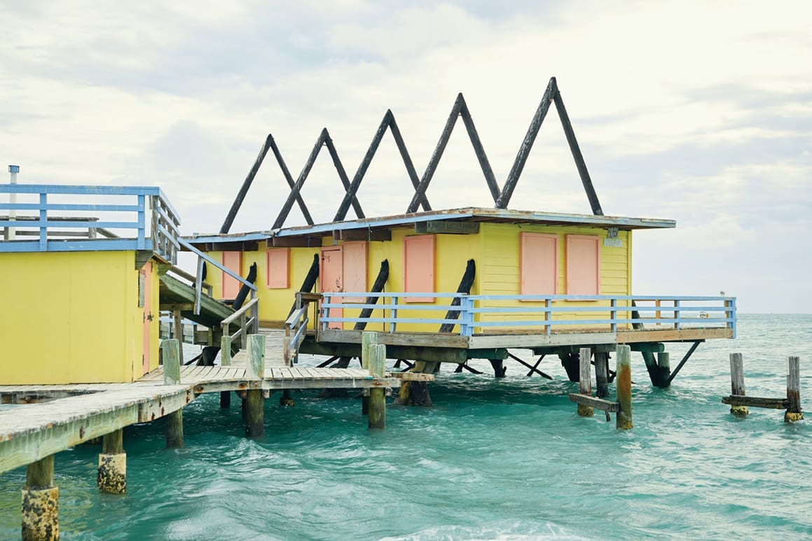 Stiltsville: Stilted, Jilted & Re-builted 4 A yellow a frame Stiltsville home with blue, coral and wooden details. Wooden triangles jut from the flat roof.