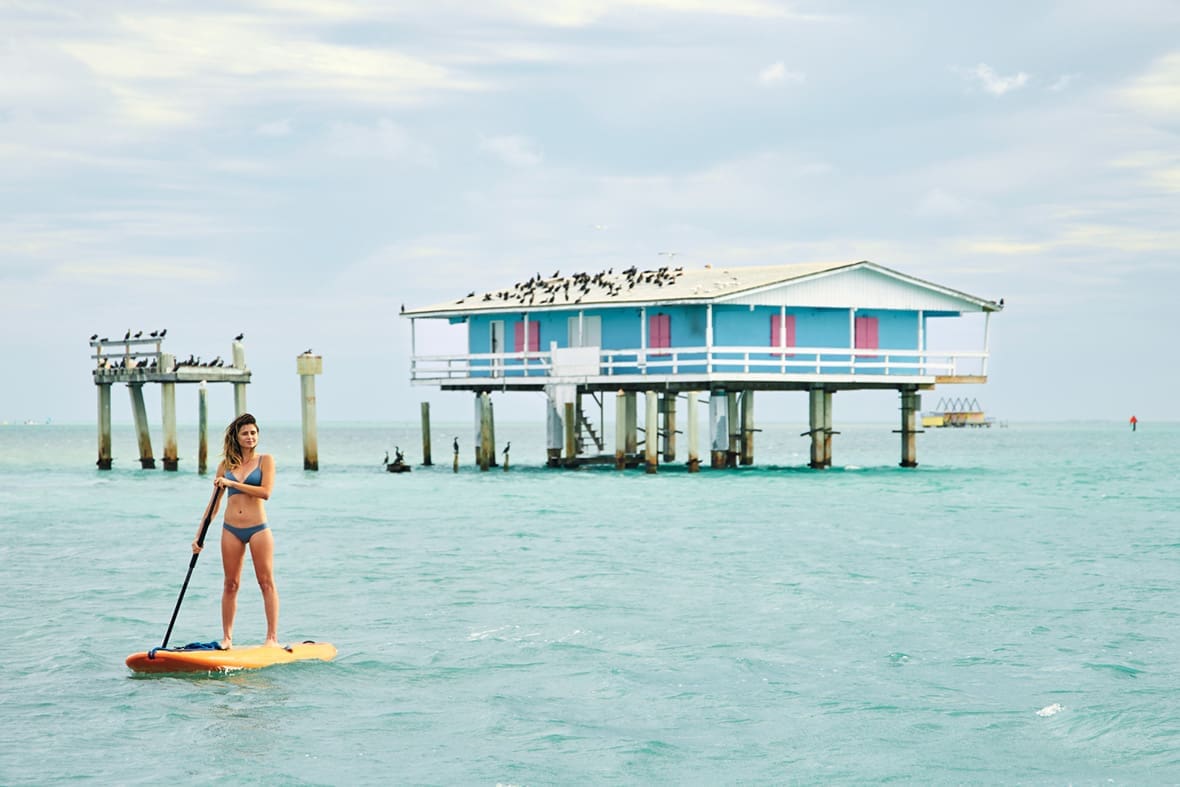 Stiltsville: Stilted, Jilted & Re-builted 6 Ashley wears a bikini and stands on a yellow paddleboard. In the background, a blue Stiltsville house. There is a flock of birds on the roof.