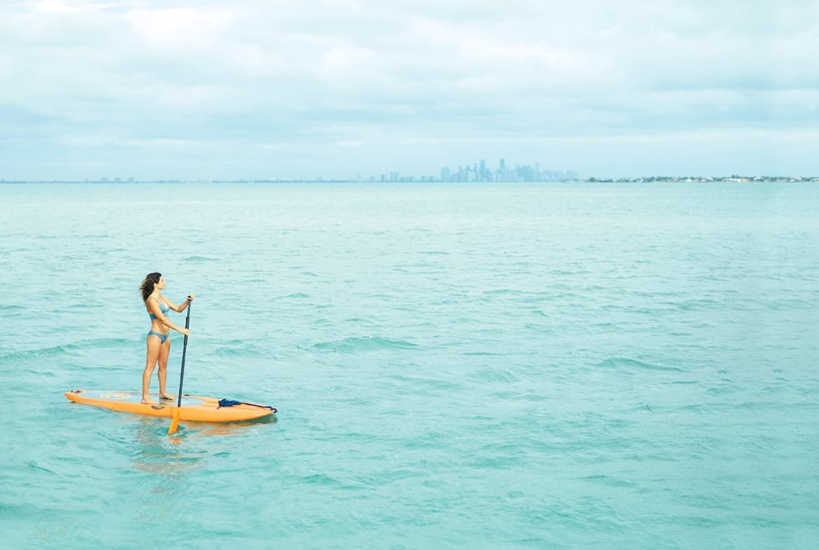 Stiltsville: Stilted, Jilted & Re-builted 2 Ashley stands on the yellow paddle board while wearing a blue bikini. She is in the bottom left corner of the photo and water fills most of the rest of the frame. A hazy Miami skyline is in the distance.