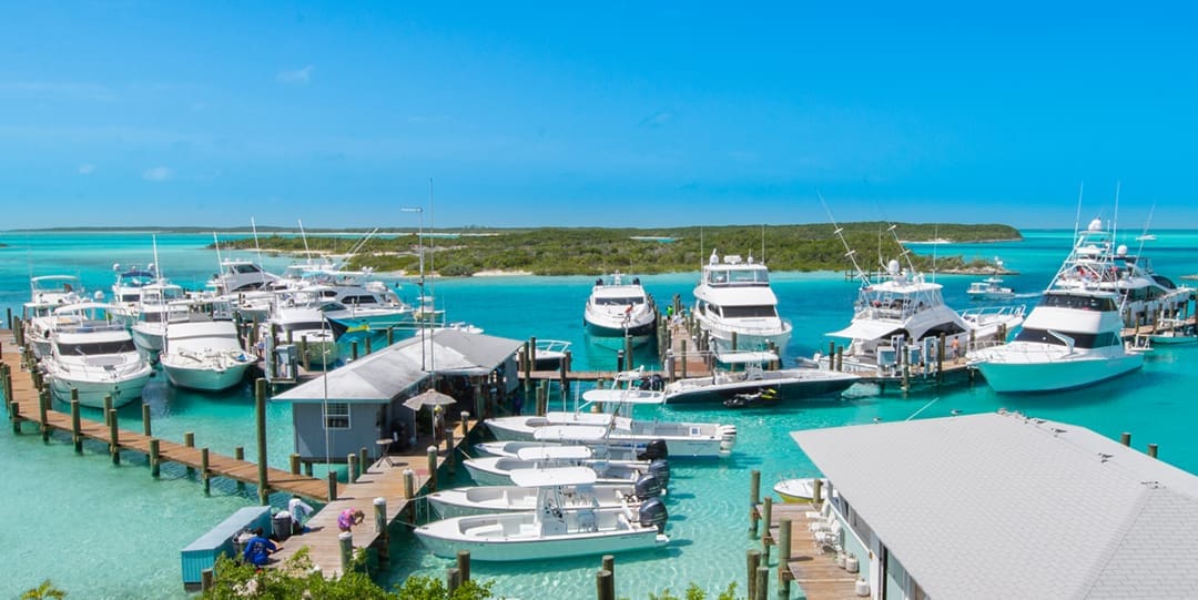 Boats of various sizes are docked in this Man-o-war Cay, an island in the Bahamas, marina. The water is bright blue and a natural, green island is in the background.
