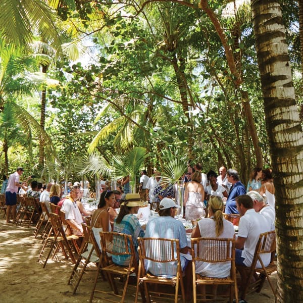 A large group gathers for a meal in the sand under the shade of tropical foliage. 