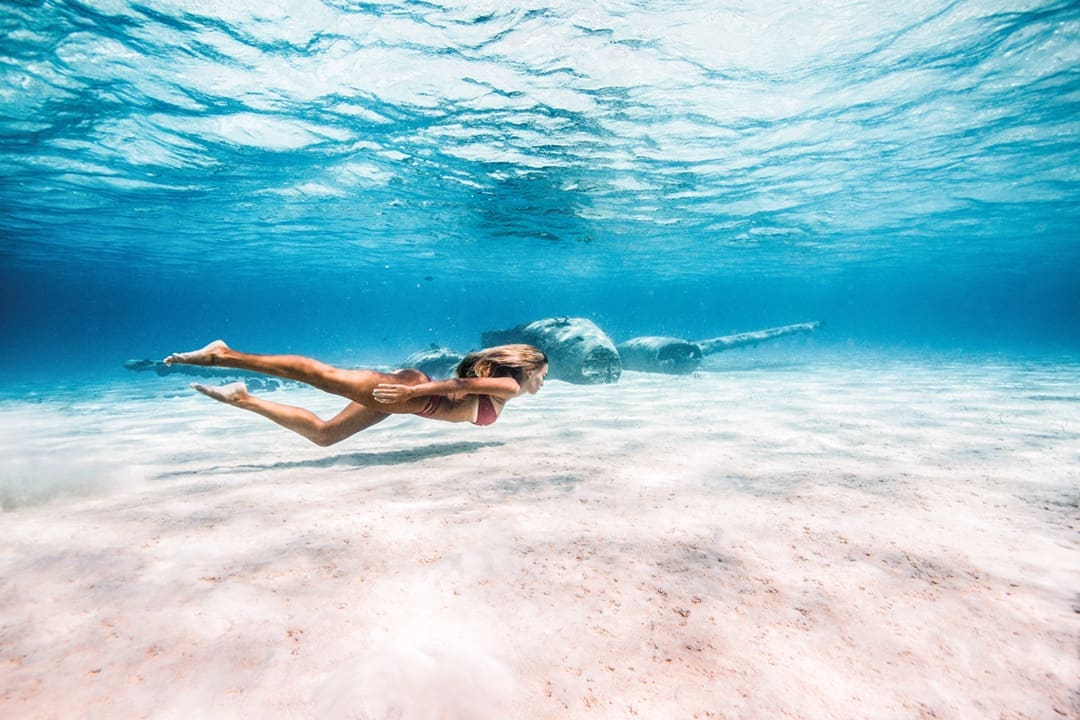 A woman snorkels in the Bahamas in front of a sunken airplane wreck in this underwater photo. Dark shapes are hard to make out but are the plane. The water is clear. 
