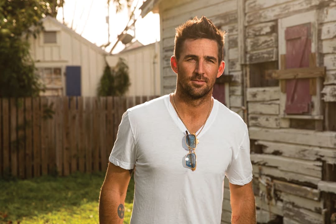 Jake Owen: The Sunshine Son 5 Jake Owen wear a white v-neck T-shirt and a neutral expression. His sunglasses are folded onto his thin necklace.
