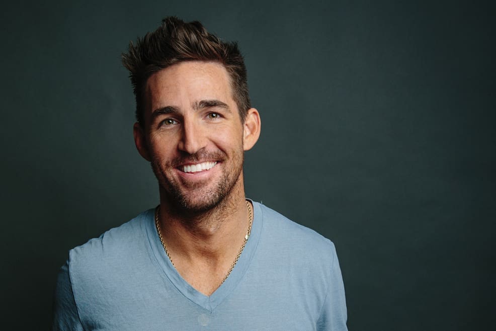 Jake Owen: The Sunshine Son 1 Jake Owen smiles in a portrait. He has light stubble and wears a blue v-neck T-shirt.