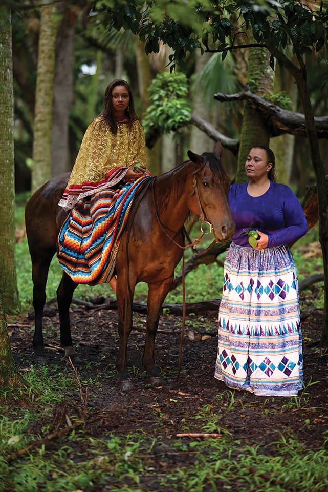 Wearing traditional Seminole dresses, Morgan and her mother Louvella Yates pick fruits on the Brighton Reservation. Morgan sits on a horse and her mother stands beside them.