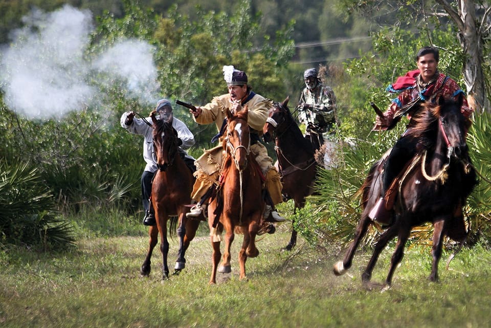 In this photo, a cloud of smoke comes out from a gun used in the re-enactment. Men ride horses and move through a natural area.