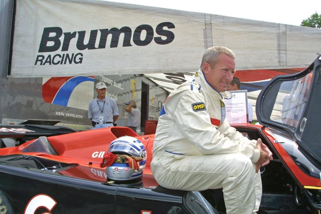 Racing Icon Hurley Haywood Gets Real About His Personal Life & Career 13 An older Haywood wears a race uniform and sits on the car. In the background, an awning reads Brumos racing.