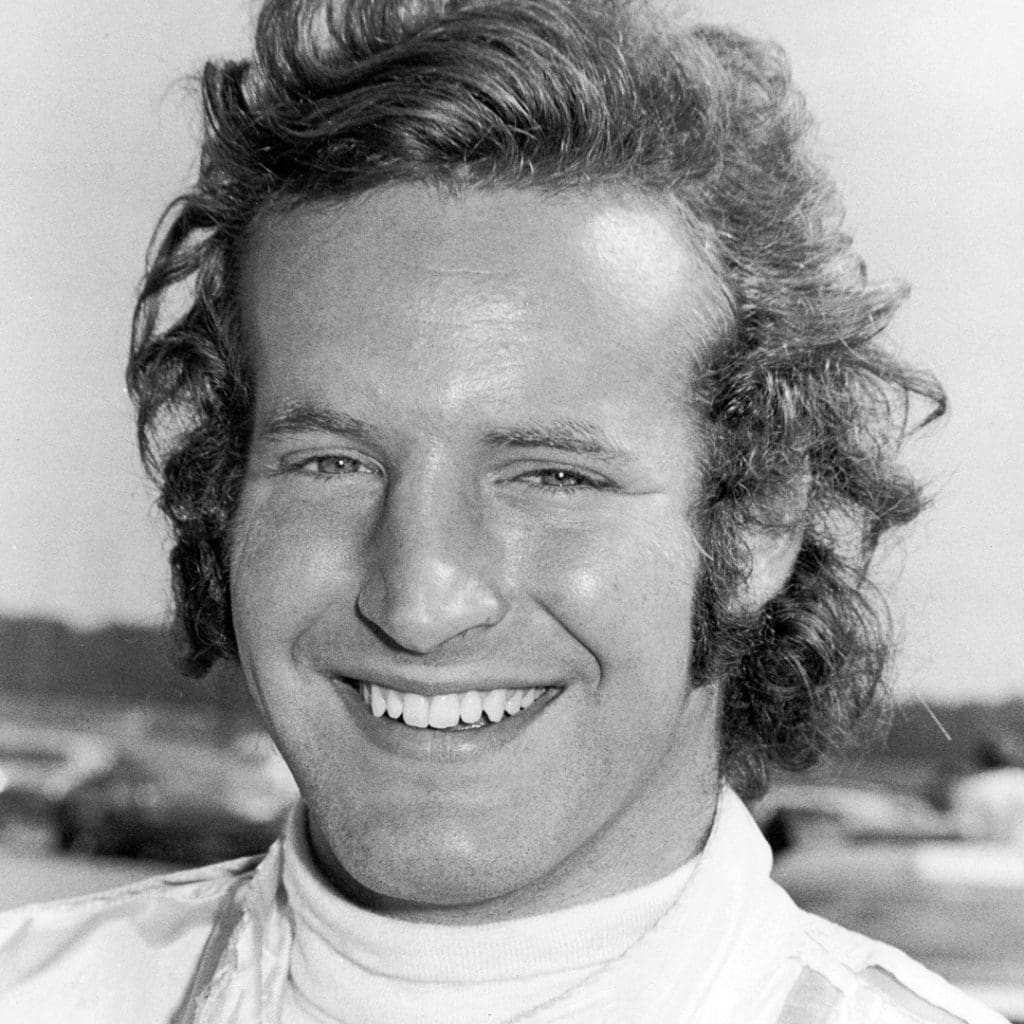 Racing Icon Hurley Haywood Gets Real About His Personal Life & Career 2 A black and white photo of young, smiling Haywood. His hair is a bit wavy and flows from his hair past his ears and a bit down his neck.