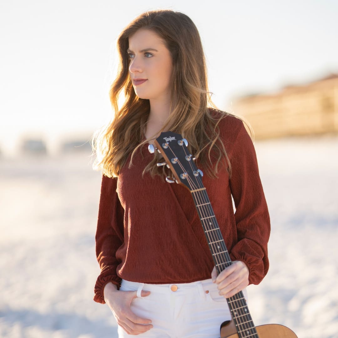 Allison Clarke stands on the beach in a rust top holding her guitar.