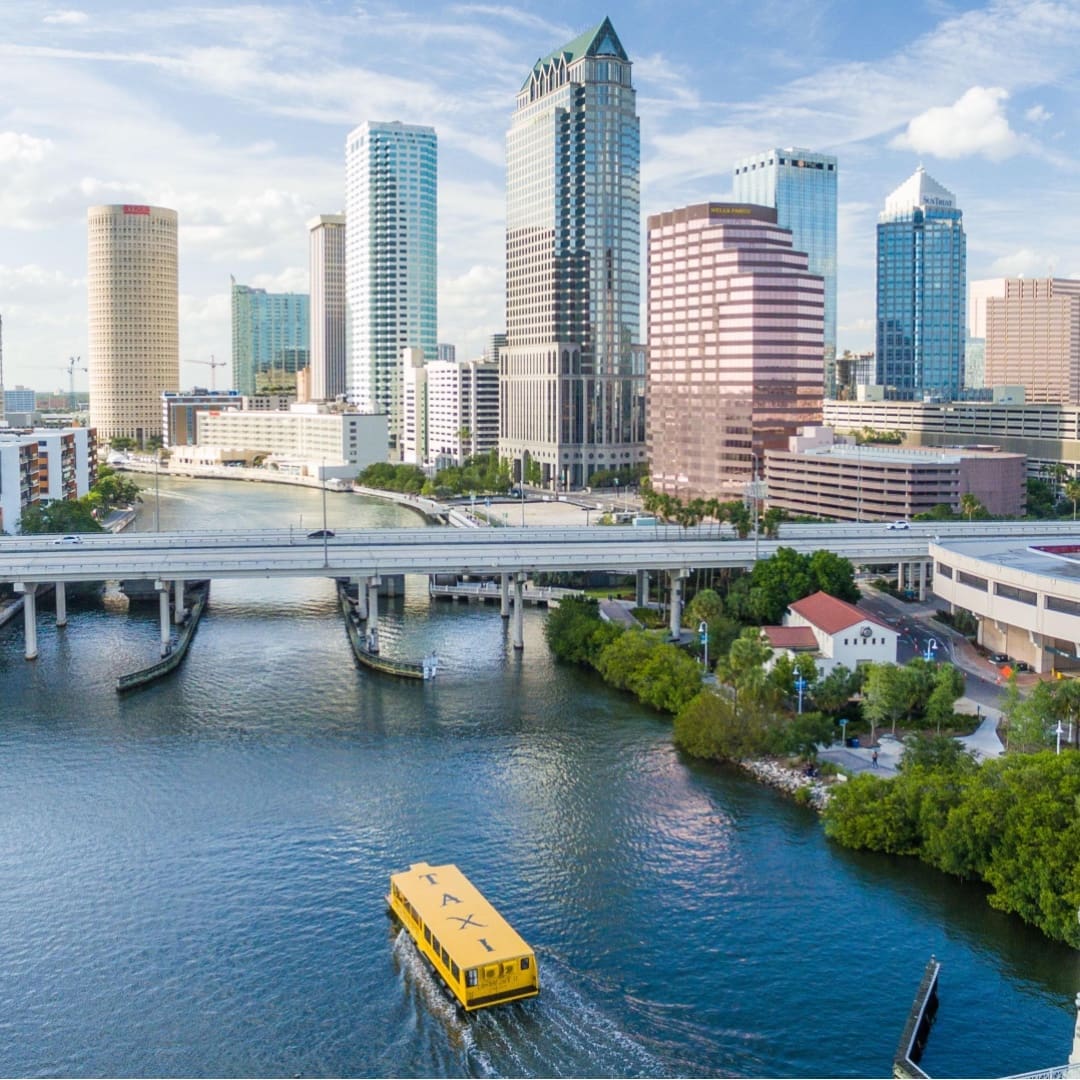 A water taxi flows through the channel of water. In the distance is a bridge for cars going over the water and the Tampa Bay skyline.