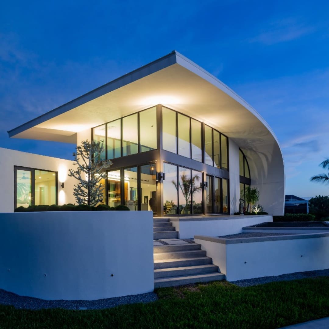 A photo of a white home with sculptural design. It is dusk and the house glows against the sky.