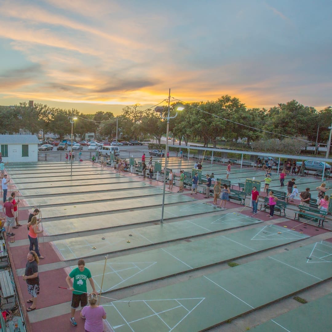 ns of players of all ages gather around the shuffleboards at the St. Petersburg Shuffleboard Club. It is sunset.