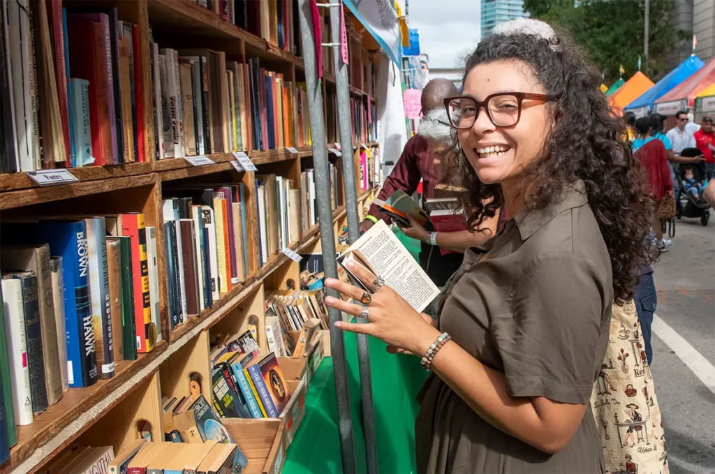 A young woman smiles while selecting a book from a packed shelf.